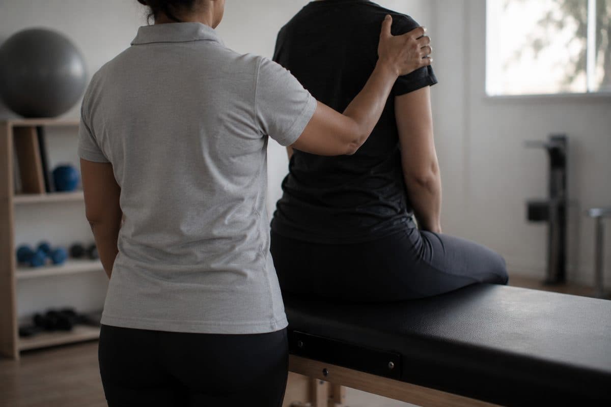Atendimento de fisioterapia numa sala clara: a profissional, de costas, apoia a mão no ombro da paciente sentada na maca