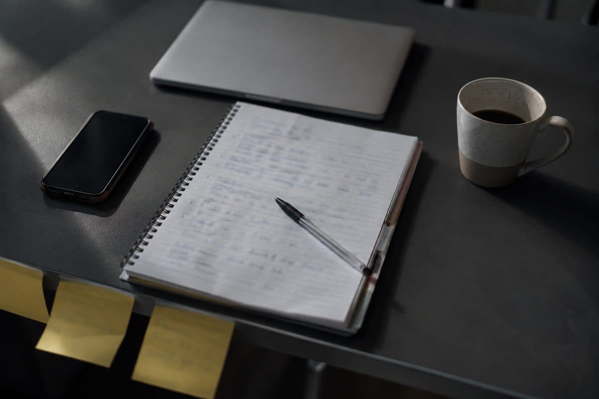 Mesa de trabalho com um caderno de anotações aberto, celular com a tela apagada, notebook fechado e post-its colados na borda